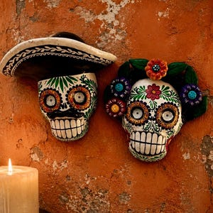 May include: Two decorative skull wall plaques featuring intricate hand-painted floral and geometric patterns in vibrant colors. One skull wears a black and white sombrero, while the other is adorned with a crown of colorful sculpted flowers.