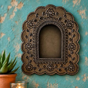 May include: An arched wall mirror with a dark bronze finish and a scalloped edge. The frame features embossed floral patterns and intricate dot detailing surrounding a central arched glass pane, creating a textured and metallic appearance.