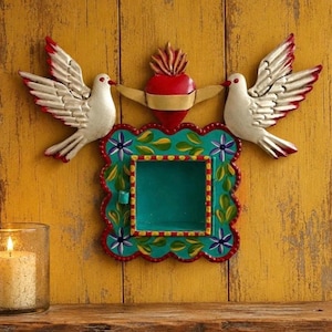 May include: A wall-mounted metal shadow box featuring a central teal square frame decorated with painted green leaves and purple flowers. Above the frame sits a red heart with a gold banner, flanked by two white doves with red-tipped wings.