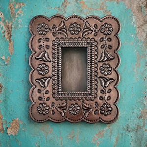 May include: A rectangular picture frame with a scalloped edge, crafted from dark metallic material. The surface features embossed floral patterns and leaf motifs surrounding a central rectangular opening with a beaded inner border.