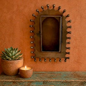 May include: A decorative brass-toned metal frame with a rectangular opening, mounted on an orange wall. A succulent in a terracotta pot and a lit candle in a small, round holder sit on a wooden surface below the frame.