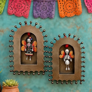 May include: Two arched, bronze-toned metal wall frames with scalloped edges, each containing a hanging ornament. One ornament features a figure with orange butterfly wings and a black dress, the other a skeleton figure with orange butterfly wings.