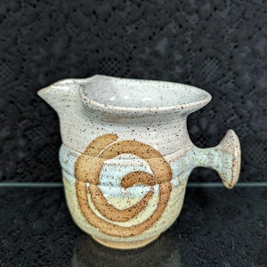 May include: A white ceramic pitcher with a brown swirl design and a handle. The pitcher has a speckled glaze and a slightly textured surface.