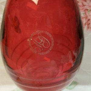 May include: A red glass vase with a clear glass base. The vase has a circular etched label that reads "HANDBLOWN ARTISANS - VINEYARD AT WESTLAKE VILLAGE - MARGIE'S GARDEN".