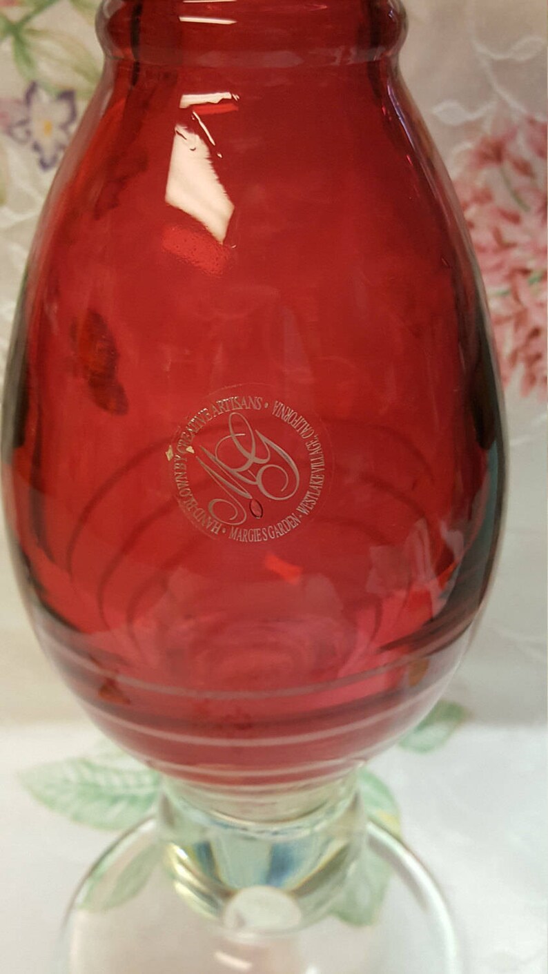 May include: A red glass vase with a clear glass base. The vase has a circular etched label that reads "HANDBLOWN ARTISANS - VINEYARD AT WESTLAKE VILLAGE - MARGIE'S GARDEN".
