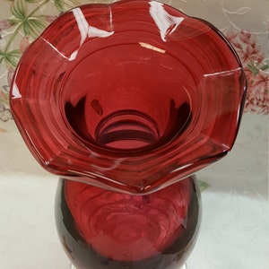 May include: A deep red glass vase with a ruffled top and a round base. The vase is made of clear glass with a deep red tint.