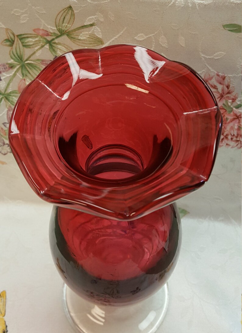 May include: A deep red glass vase with a ruffled top and a round base. The vase is made of clear glass with a deep red tint.