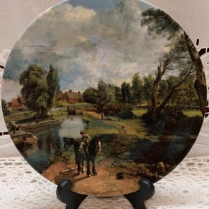 May include: A ceramic plate with a painting of a rural landscape with a river, a bridge, and a horse-drawn cart. The painting is framed by a white border and the plate is on a black stand.