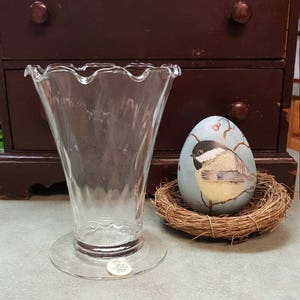 May include: A clear glass vase with a ruffled rim and a wide base. The vase is sitting on a white surface. The vase is made by the company "C. Spencer Gifts, Inc."
