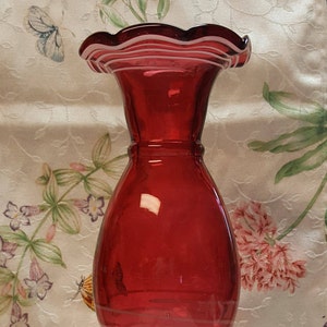May include: A red glass vase with white ruffled trim and white stripes. The vase has a round body and a narrow neck.