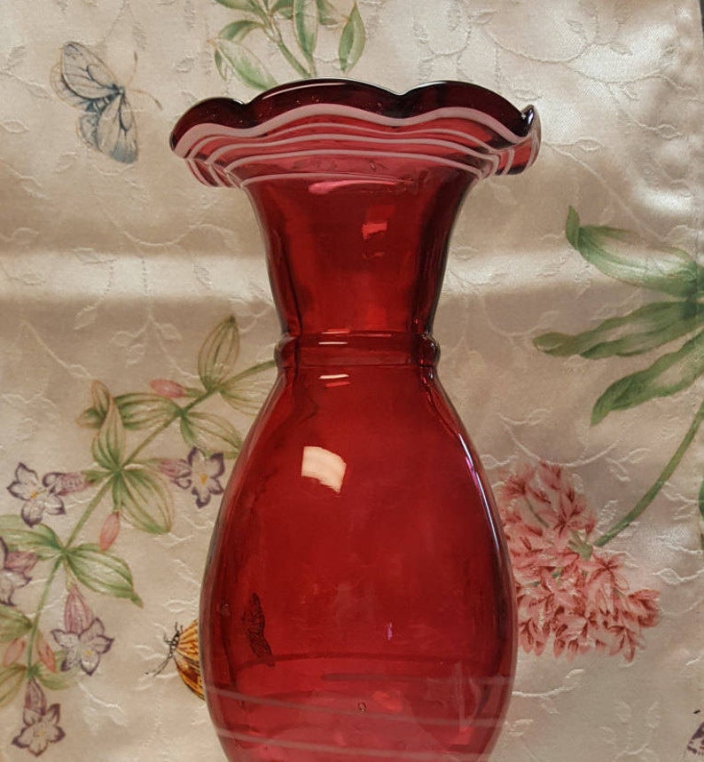 May include: A red glass vase with white ruffled trim and white stripes. The vase has a round body and a narrow neck.