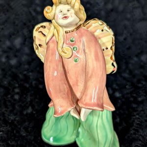 May include: A ceramic angel figurine with a peach-colored robe and green shoes. The angel has blonde curly hair and gold-trimmed wings. Three green gems adorn the front of the robe. The figurine is a decorative item.