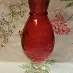 May include: A red glass vase with a ruffled top and a clear glass base. The vase has a swirl design and is perfect for displaying flowers.