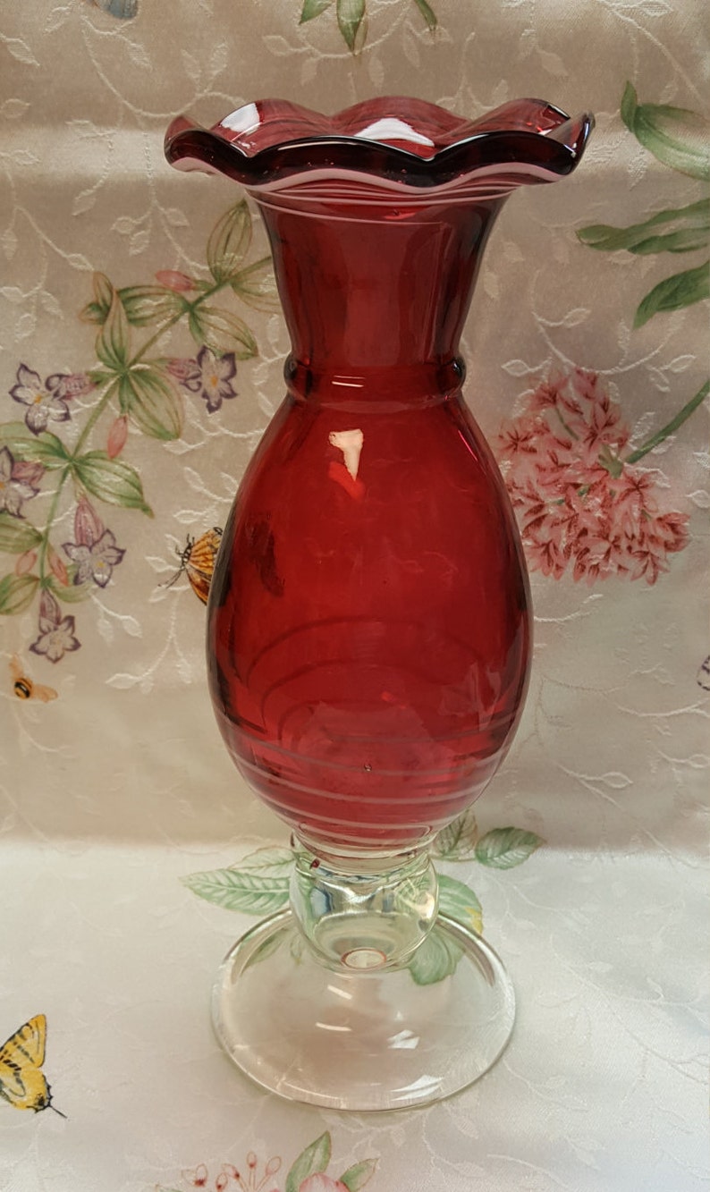 May include: A red glass vase with a ruffled top and a clear glass base. The vase has a swirl design and is perfect for displaying flowers.