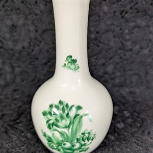 May include: A white ceramic vase with a gold rim and a bulbous base. The vase is decorated with green floral designs. The vase is approximately 7 inches tall.