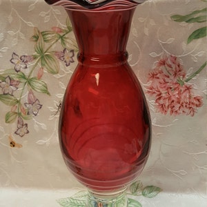 May include: A red glass vase with a ruffled top and a clear glass base. The vase has a textured, ribbed design.