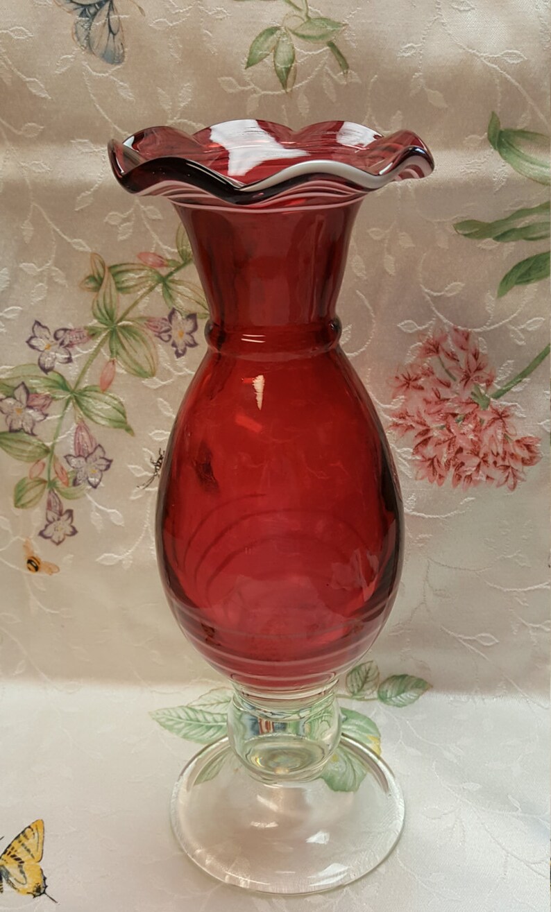 May include: A red glass vase with a ruffled top and a clear glass base. The vase has a textured, ribbed design.