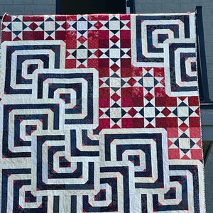 May include: A handmade quilt with a geometric design in red, white, and navy blue. The quilt features a repeating pattern of interlocking squares and rectangles, with a mix of solid and patterned fabrics. The quilt is hanging outside.