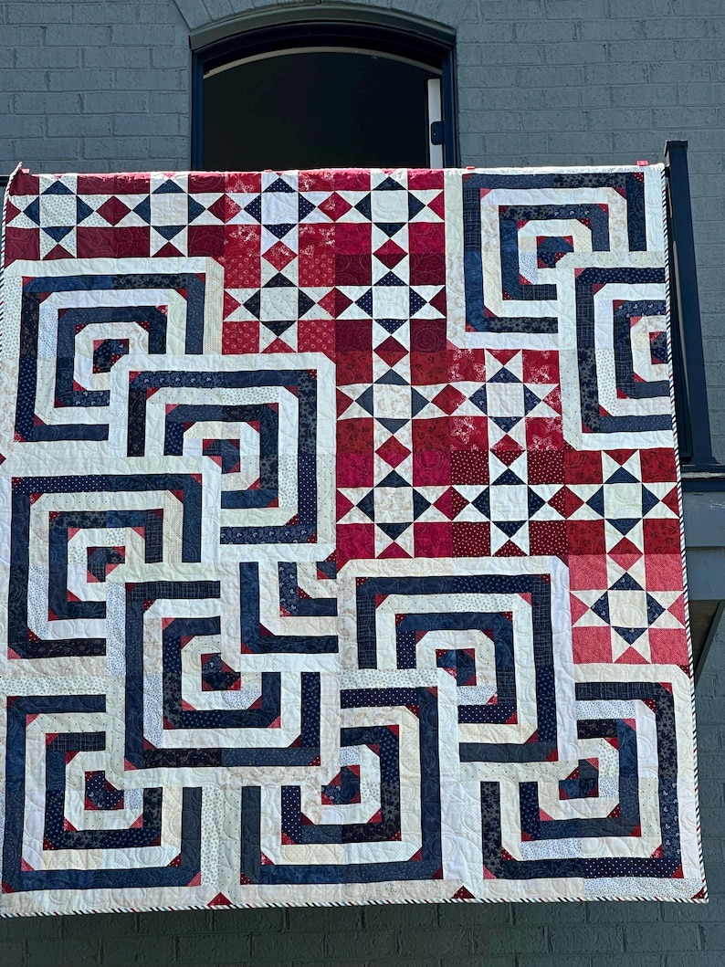 May include: A handmade quilt with a geometric design in red, white, and navy blue. The quilt features a repeating pattern of interlocking squares and rectangles, with a mix of solid and patterned fabrics. The quilt is hanging outside.