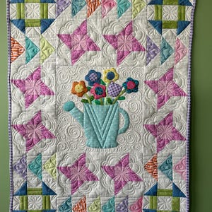 May include: A handmade quilt with a floral theme. The quilt features a light blue watering can with colorful fabric flowers. Pink star-shaped designs and geometric patterns in various colours are arranged on a white background. The quilt is bordered with a purple and white gingham fabric.