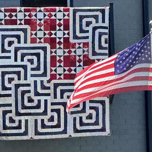 May include: A red, white, and blue quilt with a geometric design hangs on a grey building. The quilt features a repeating pattern of squares and rectangles. An American flag waves in the wind next to the quilt.