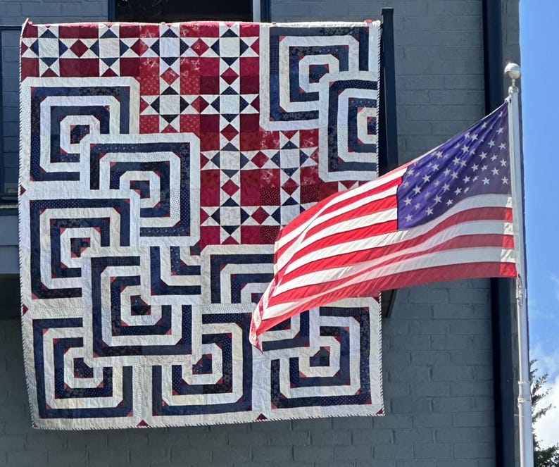 May include: A red, white, and blue quilt with a geometric design hangs on a grey building. The quilt features a repeating pattern of squares and rectangles. An American flag waves in the wind next to the quilt.