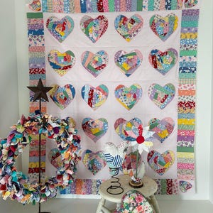 Op de afbeelding: Een handgemaakte quilt met geappliceerde harten in verschillende kleurrijke stofpatronen. De quilt wordt getoond met een bijpassend hartvormig kussen, een stoffen krans en andere decoratieve items. De quilt is een patchwork ontwerp met een roze achtergrond.