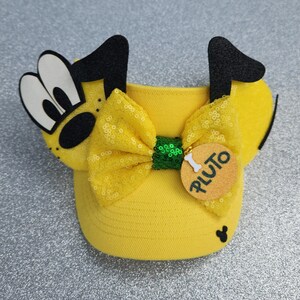 May include: A yellow visor featuring a Pluto character design with black ears, a yellow sequined bow, and a green sequined center. A round tan tag hangs from the bow with a white bone icon and the text Pluto printed in black. A small black Mickey icon is on the brim.