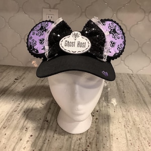 May include: Black baseball cap with mouse ears. The ears are purple with a black filigree design. The center of the cap has a black sequined bow with a white label that reads "Ghost Host". A purple heart is on the side of the cap.
