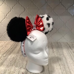 May include: A headband featuring two large fuzzy ears, one solid black and one white with black spots. A shiny metallic red bow with a silver rhinestone center sits between the ears. The headband band is white with black polka dots and a Disney tag.