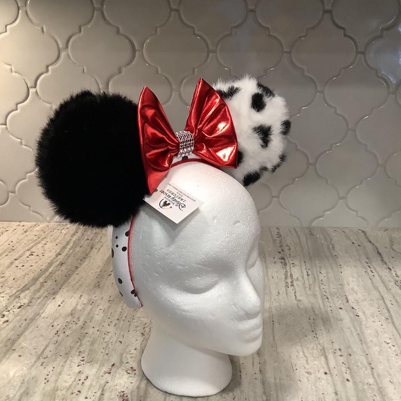 May include: A headband featuring two large fuzzy ears, one solid black and one white with black spots. A shiny metallic red bow with a silver rhinestone center sits between the ears. The headband band is white with black polka dots and a Disney tag.