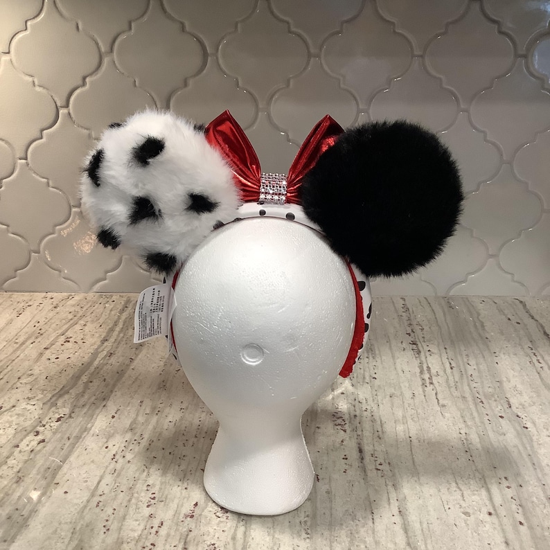 May include: A headband featuring two large fluffy ears, one white with black spots and one solid black. A shiny red metallic bow sits between the ears, accented with a central strip of clear rhinestones. The headband band has a white and black pattern.