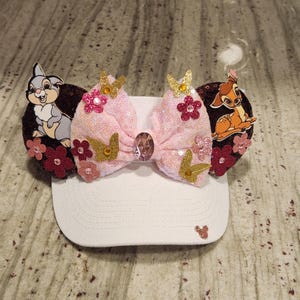 May include: White baseball cap with a pink sequined bow, adorned with gold butterflies, pink flowers, and a large gem. Features Bambi and Thumper character cutouts on each side.