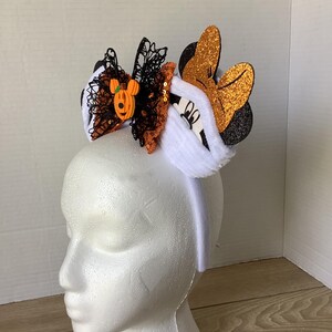 Op de afbeelding: Een wit hoofdband met pluche witte oren en een zwart-oranje strik met een pompoen Mickey Mouse-bedel. De strik is versierd met zwarte kant en oranje pailletten.