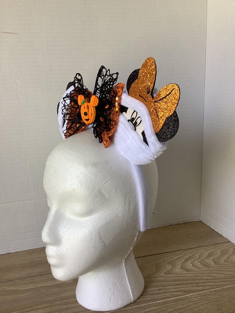 Op de afbeelding: Een wit hoofdband met pluche witte oren en een zwart-oranje strik met een pompoen Mickey Mouse-bedel. De strik is versierd met zwarte kant en oranje pailletten.