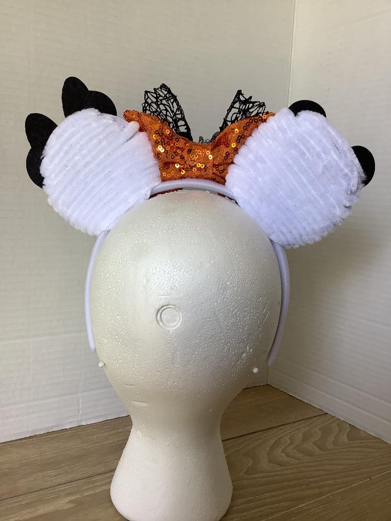 Op de afbeelding: Witte, pluche muizenoren met een oranje en zwarte pailletten strik. De strik heeft zwarte kant accenten.