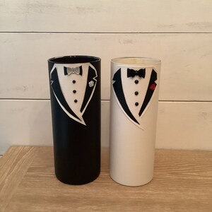 May include: Two cylindrical vases decorated with tuxedo designs. The vase on the left is black with a silver bow tie and the vase on the right is white with a black bow tie and a red flower.