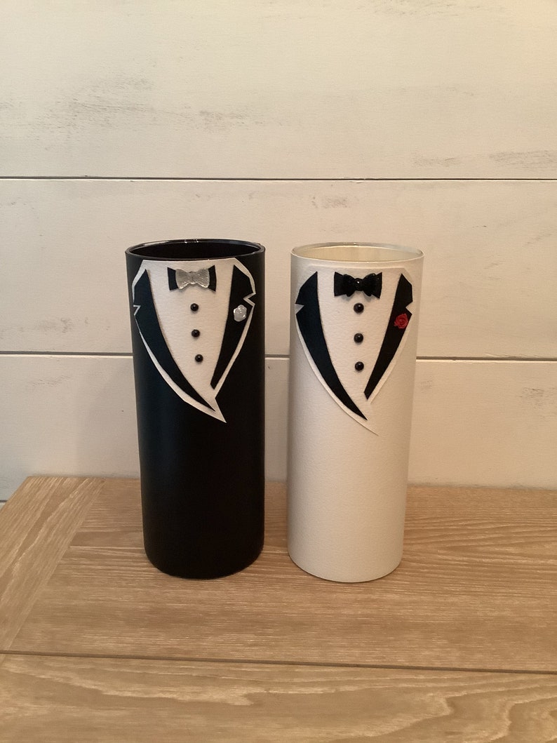 May include: Two cylindrical vases decorated with tuxedo designs. The vase on the left is black with a silver bow tie and the vase on the right is white with a black bow tie and a red flower.