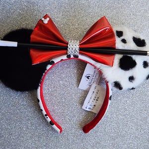 May include: A headband featuring one solid black plush ear and one white plush ear with black spots. A shiny red bow with a central band of rhinestones sits between the ears. A black and white prop cigarette holder is attached across the bow.