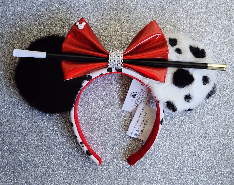 May include: A headband featuring one solid black plush ear and one white plush ear with black spots. A shiny red bow with a central band of rhinestones sits between the ears. A black and white prop cigarette holder is attached across the bow.