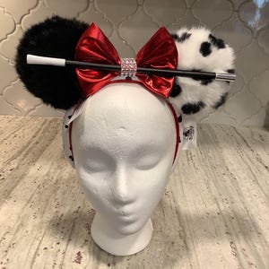 May include: A headband featuring two round ears, one solid black faux fur and one white faux fur with black spots. A shiny red metallic bow sits in the center with a rhinestone band. A long black and white cigarette holder prop is attached.