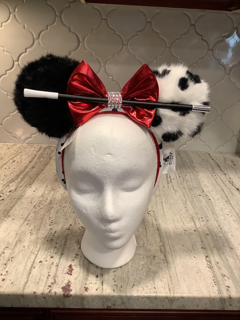 May include: A headband featuring two round ears, one solid black faux fur and one white faux fur with black spots. A shiny red metallic bow sits in the center with a rhinestone band. A long black and white cigarette holder prop is attached.