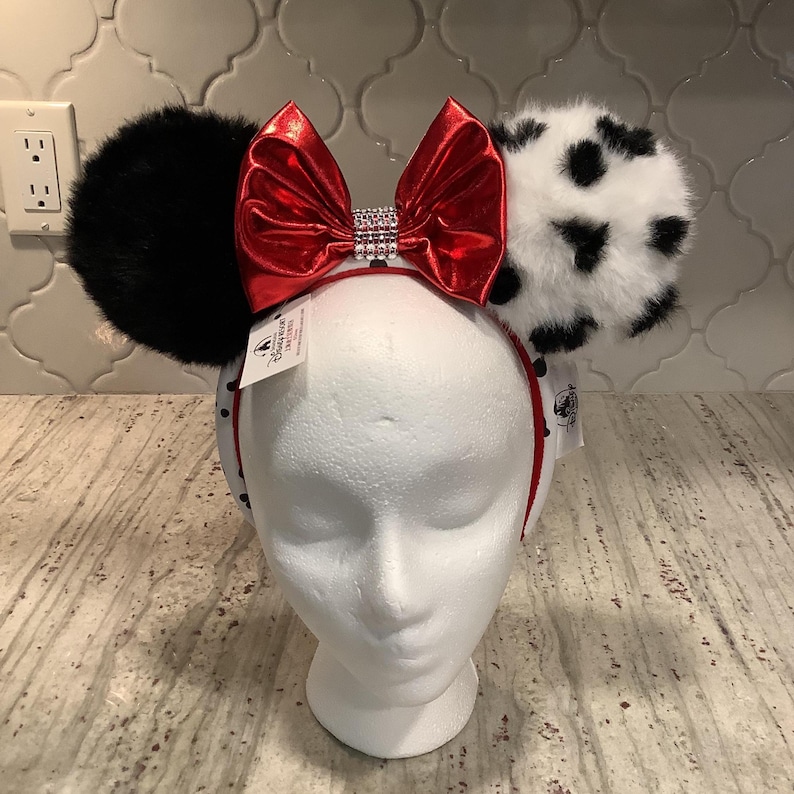 May include: A headband featuring two large ears made of faux fur, with the left ear in solid black and the right ear in white with black spots. A shiny metallic red bow with a central band of clear rhinestones sits between the ears.
