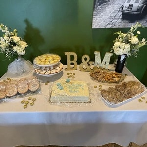 May include: A white table with a white tablecloth is set up for a wedding reception. There are various desserts on the table, including cookies, cake, and pastries. The table is decorated with gold hearts and the letters "B&M" in gold. There are also two bouquets of white flowers in vases.
