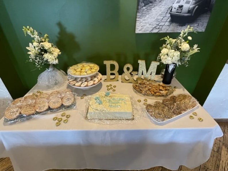 May include: A white table with a white tablecloth is set up for a wedding reception. There are various desserts on the table, including cookies, cake, and pastries. The table is decorated with gold hearts and the letters "B&M" in gold. There are also two bouquets of white flowers in vases.