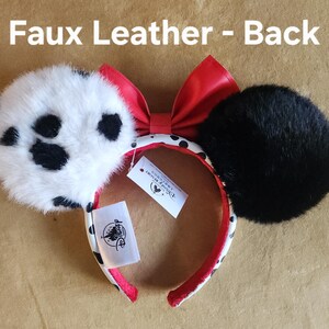 May include: A headband with two circular ears, one white with black spots and one solid black, both covered in faux fur. A red faux leather bow sits between the ears. The headband band is white with black spots and red lining. Text reads: Faux Leather - Back.