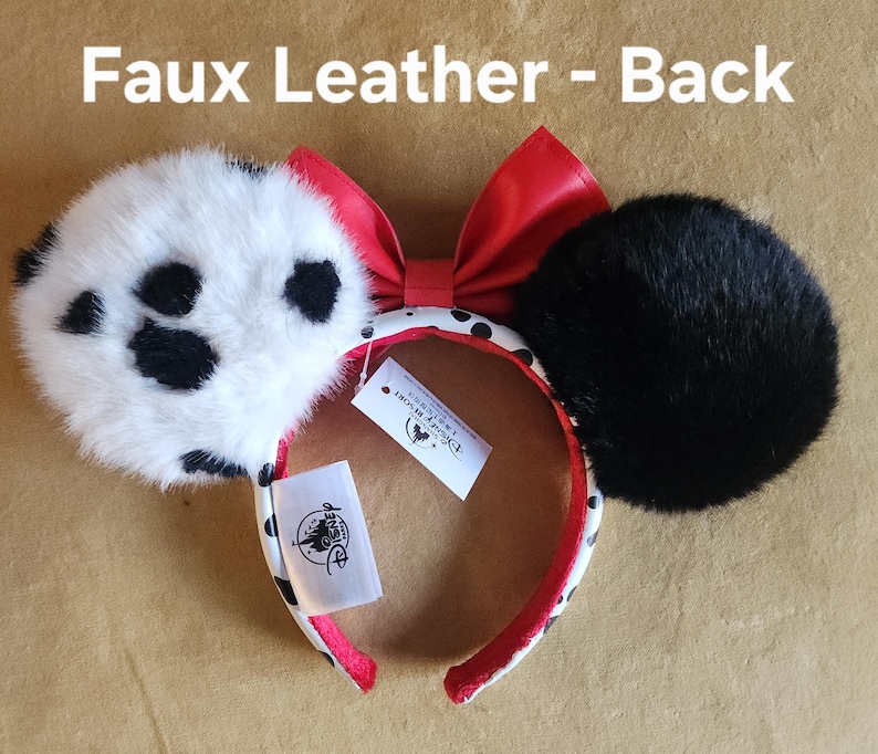 May include: A headband with two circular ears, one white with black spots and one solid black, both covered in faux fur. A red faux leather bow sits between the ears. The headband band is white with black spots and red lining. Text reads: Faux Leather - Back.