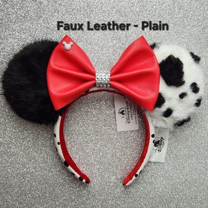 May include: Headband with two ears, one solid black faux fur and one white faux fur with black spots. A large red faux leather bow sits in the center with a silver rhinestone band and a small silver Mickey icon. Text reads Faux Leather - Plain.