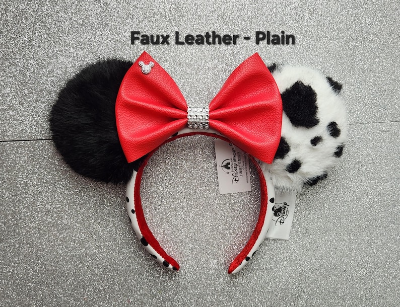 May include: Headband with two ears, one solid black faux fur and one white faux fur with black spots. A large red faux leather bow sits in the center with a silver rhinestone band and a small silver Mickey icon. Text reads Faux Leather - Plain.
