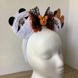 Op de afbeelding: Een wit hoofdband met een zwart en oranje strikje, een wit, pluche Mickey Mouse-oor met een mummie en een oranje Mickey Mouse-oor met een pompoen erop.